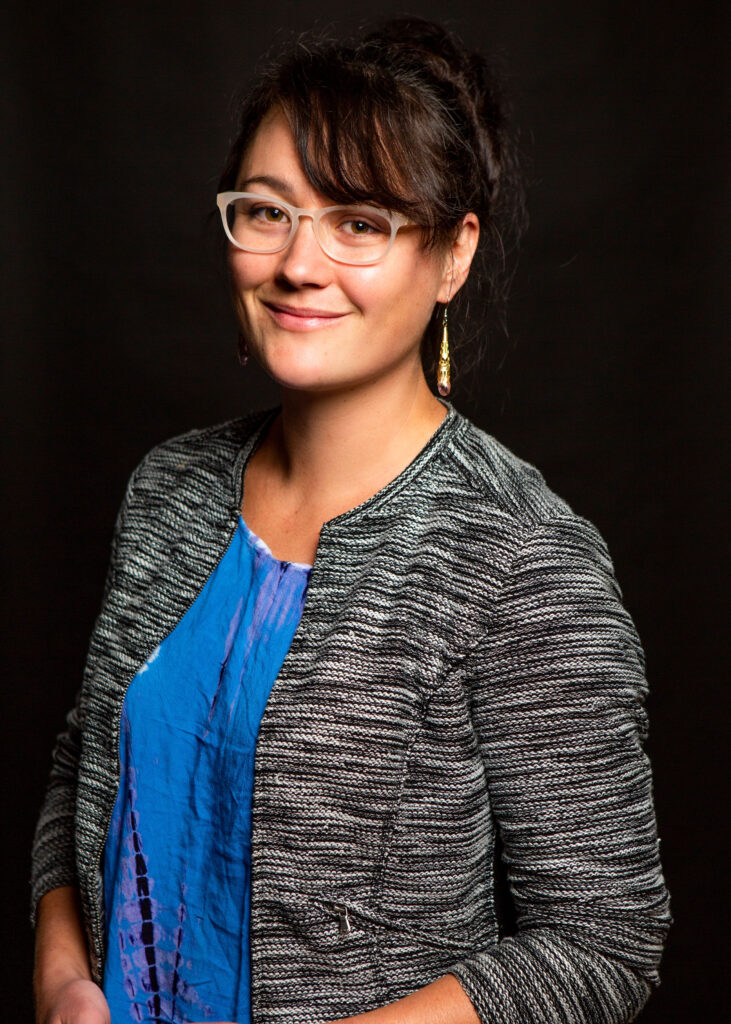 A smiling young mixed-race woman with black hair, glasses, and long dangling earrings, wearing a striped sweater and a blue shirt.