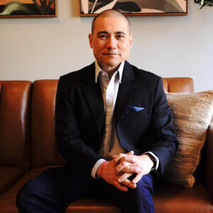 An asian man wearing a suit sits on a brown couch with his hands on his knee, smiling.