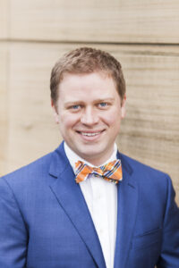 White man with short brown hair wearing a bow tie and a blue jacket with a white shirt standing in front of a beige wall