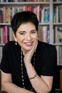 A white woman with short dark hair sits in front of a bookcase, hand on chin. Author Karen Kirsten.