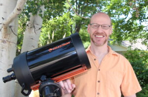White man wearing a tan shirt stands next to a tree and a telescope